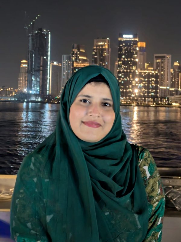 Nadiya Qudsi smiling, in a green hijab, with the Dubai skyline lit up behind her at night